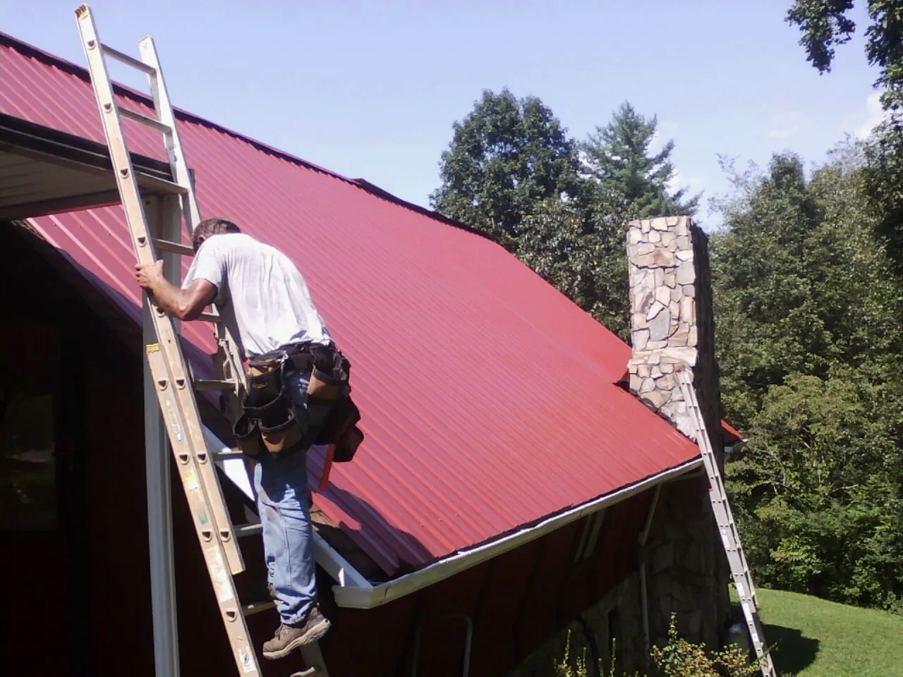Roofer on a ladder beside a red metal roof and stone chimney — Soffit & Fascia Repair by Roofing Brothers in Madison Lake